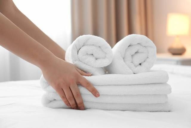 Hands arranging rolled white towels on a neatly made bed in a softly lit room.